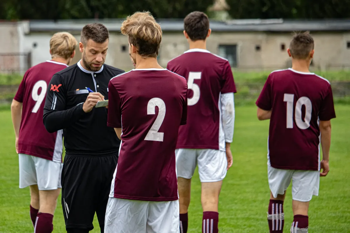 Soccer referee taking notes as team gathers on the field for a match outdoors.