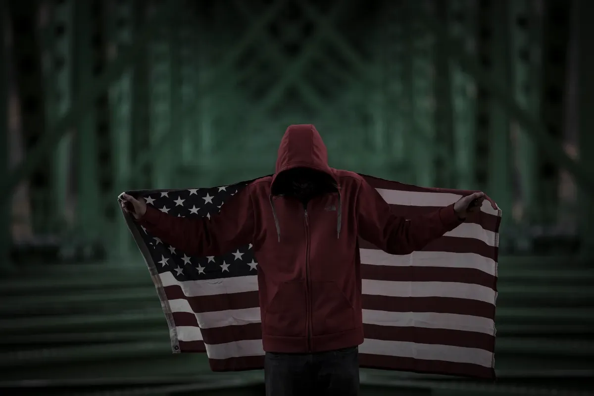 A person in a hoodie holds an American flag under a dark, moody bridge.