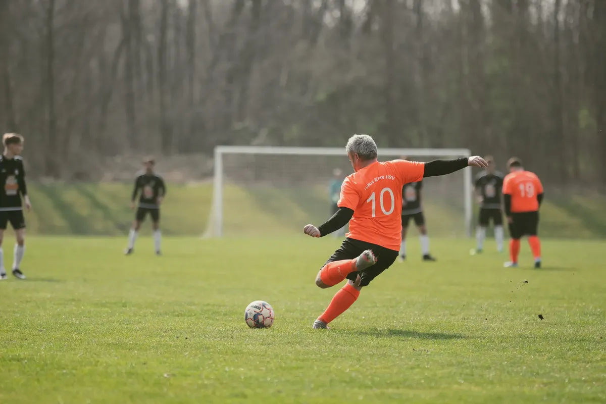 An adult soccer player in an orange jersey kicks the ball during an outdoor match on a grass field.